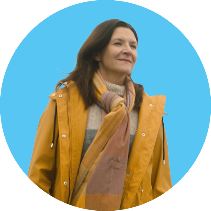 Dynes mewn cot a sgarff melyn yn edrych i'r pellter a gwenu gyda chefndir glas cylch